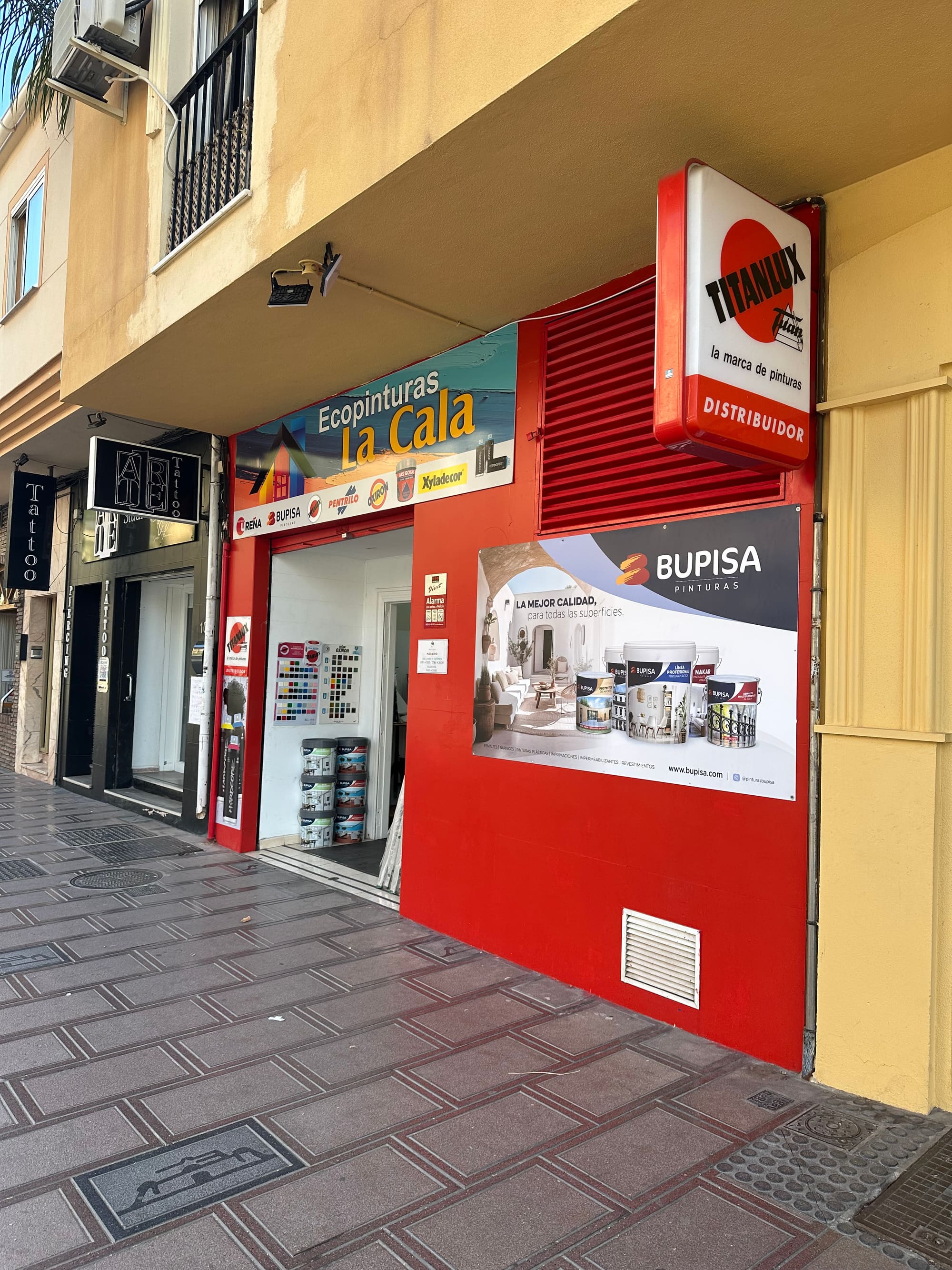 Fachada roja de Ecopinturas La Cala vista desde la acera de la Avenida de Málaga, con el rótulo de la tienda y el cartel de Titanlux.