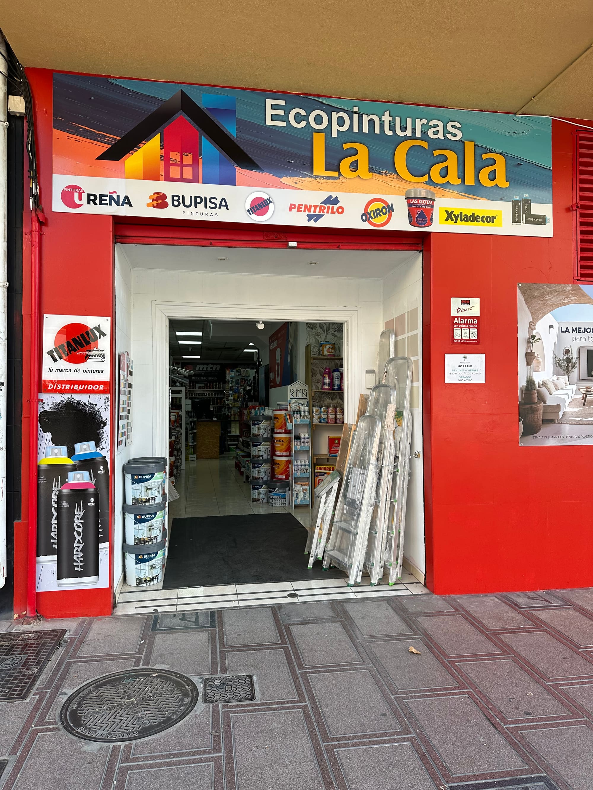 Vista frontal de la puerta de Ecopinturas La Cala abierta, con el pasillo de la tienda al fondo y varias escaleras apoyadas en la jamba.