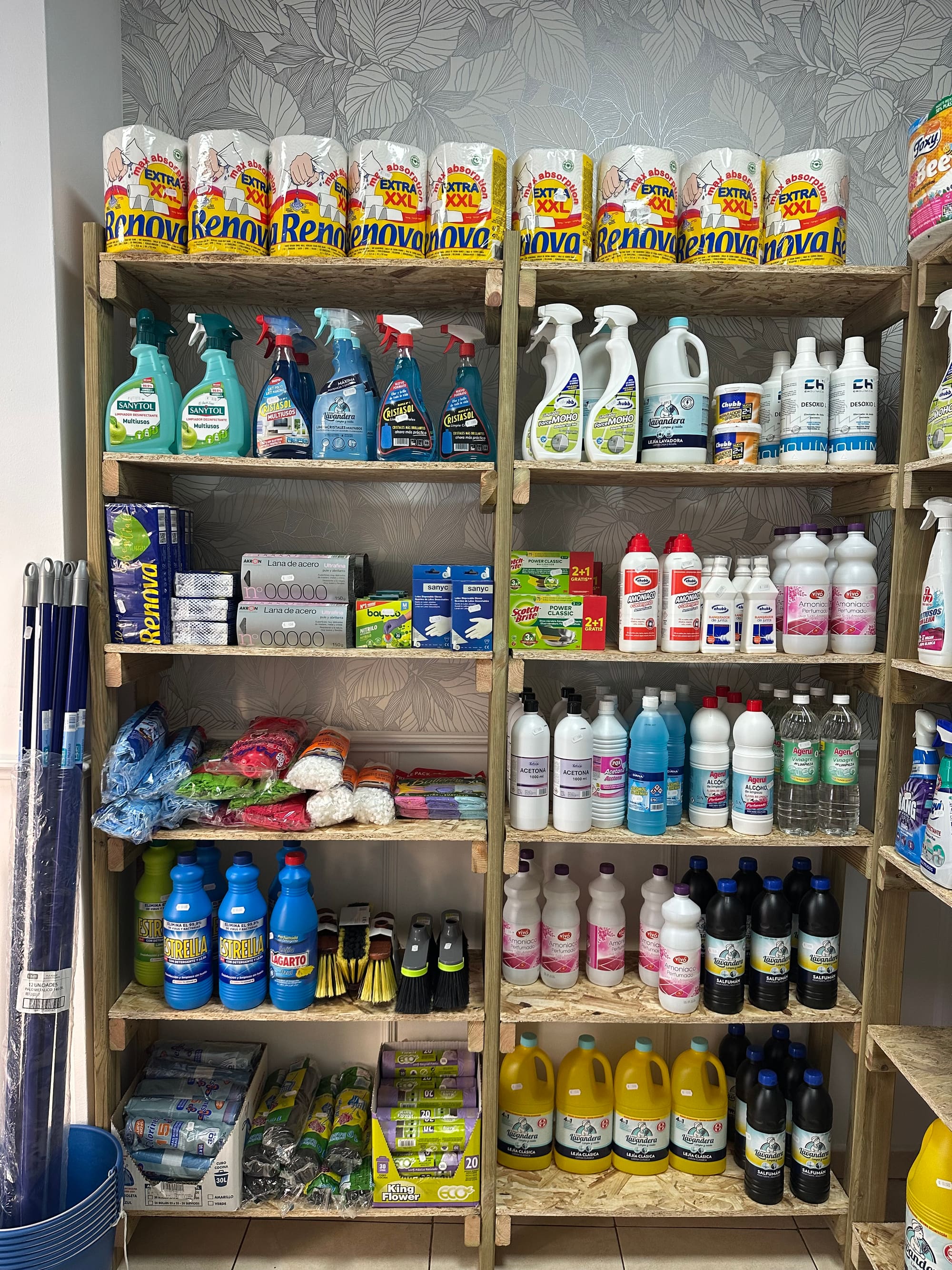 Estantería de droguería con papel de cocina Renova arriba, sprays limpiadores en el centro y garrafas de lejía abajo.