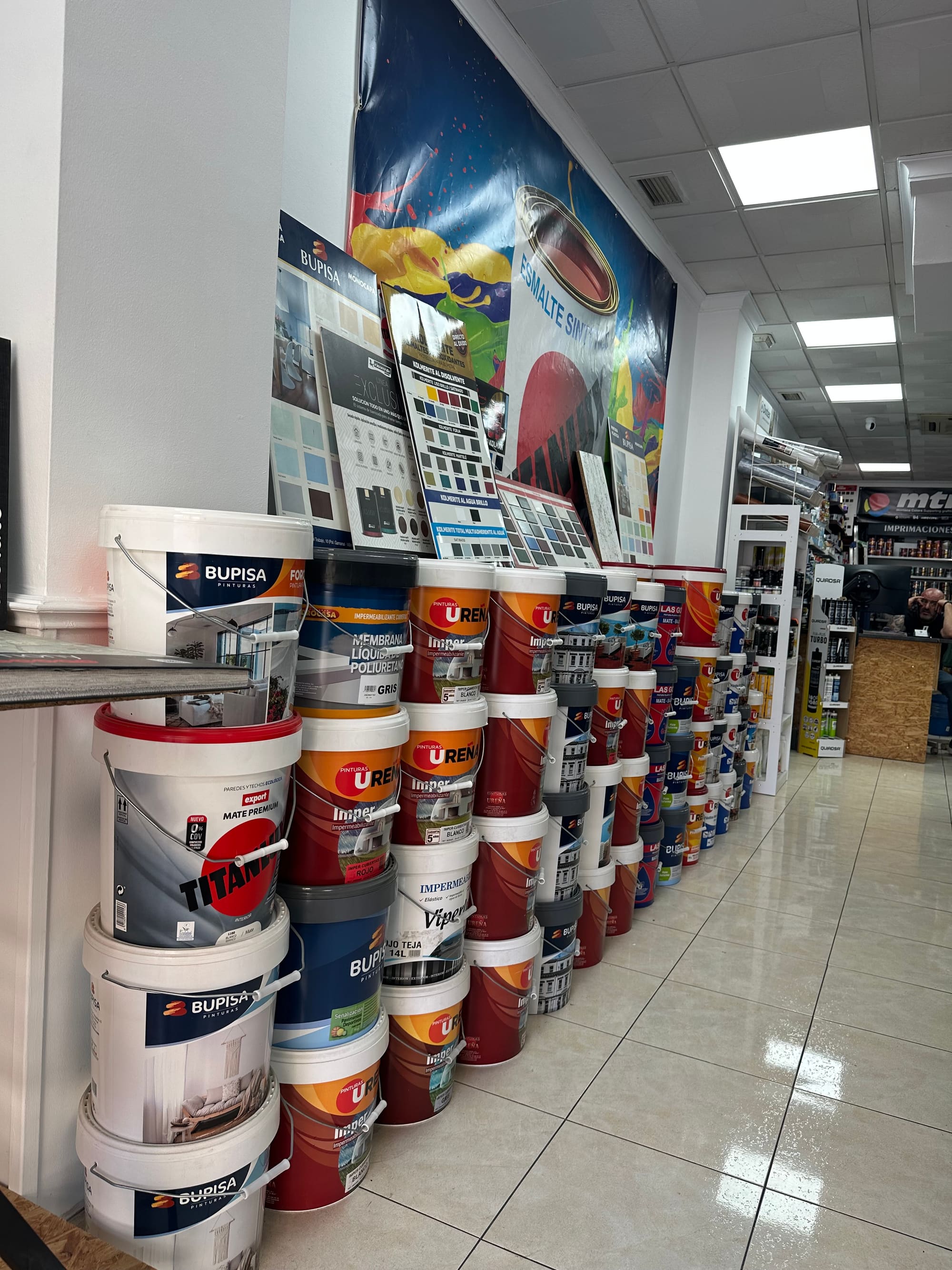 Pasillo de la tienda con cubos de pintura Bupisa, Ureña y Titan apilados a lo largo de la pared bajo un cartel de esmalte sintético.