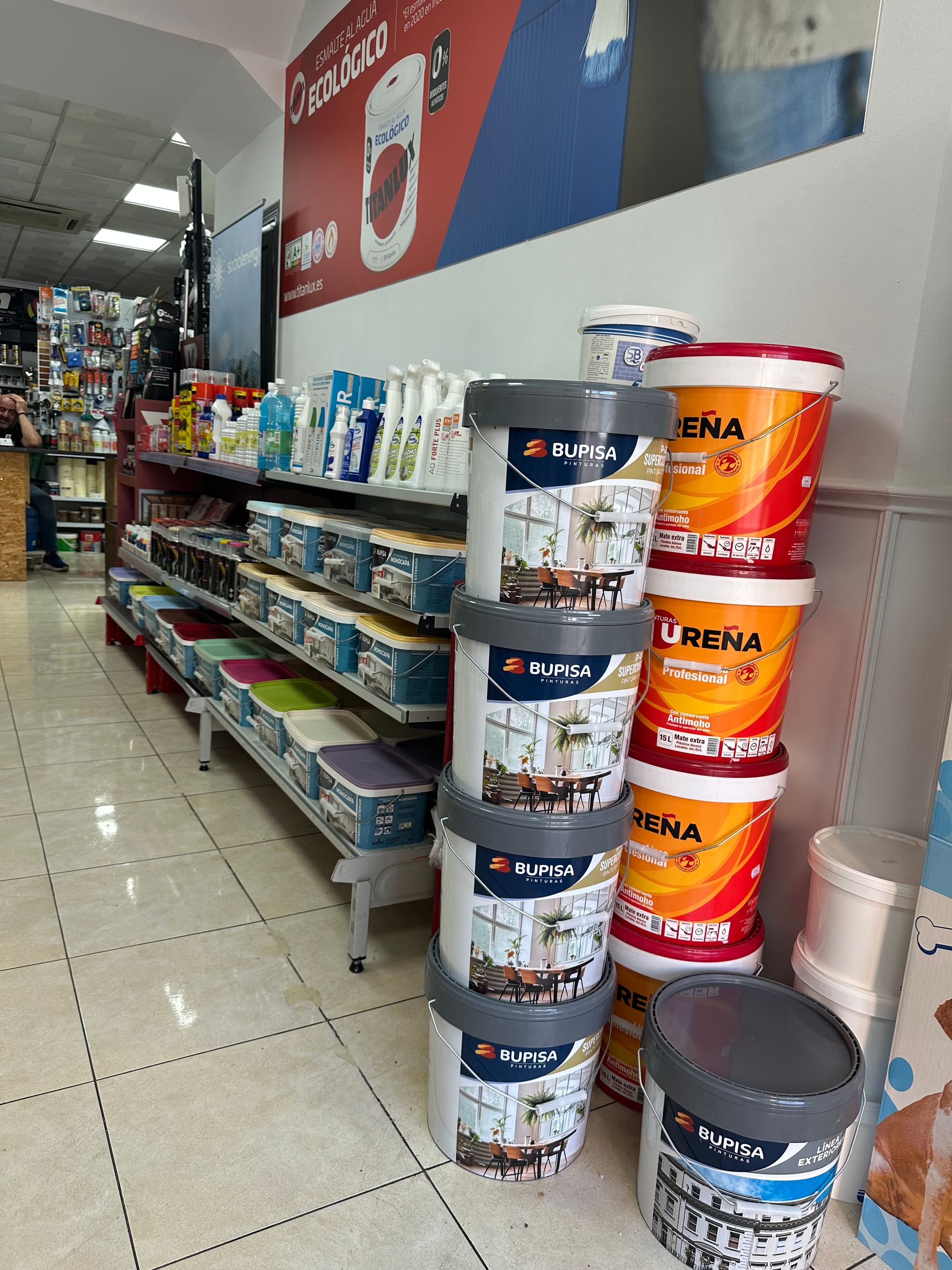 Cubos de pintura plástica Bupisa y Ureña apilados junto a una estantería con cajas de plástico de colores.